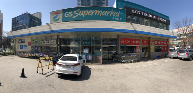 Ein Auto steht vor einem 'GS Supermarkt'-Geschäft mit anderen Fahrzeugen, Verkehrskegeln und verstreuten Gegenständen auf der Straße; Gebäude, Bäume und ein klarer blauer Himmel im Hintergrund.