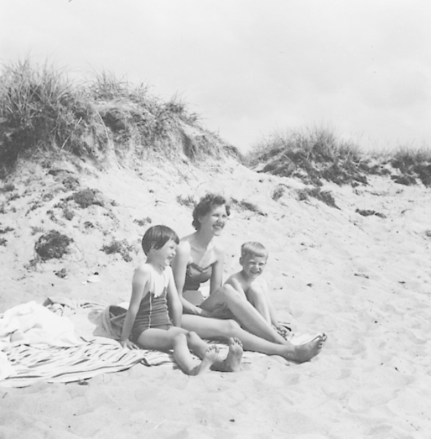Schwarz-weiß-Foto einer Frau in einem Bikini und zweier Kinder in Badebekleidung, die auf einer Decke am Strand sitzen, mit Sand, Gras und Himmel im Hintergrund.