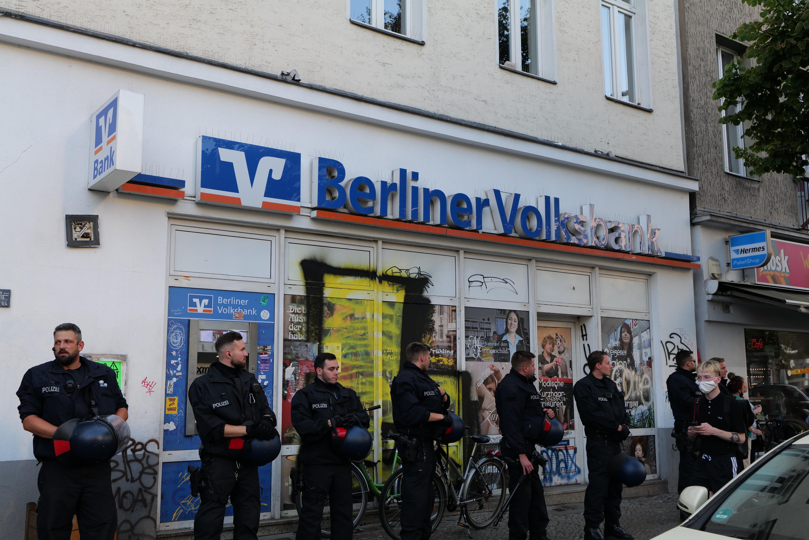 Polizisten vor einer Berliner Volksbank mit Helmen in den Händen und einem Auto auf der rechten Seite, einem Baum auf der linken Seite, Gebäude und Texttafeln im Hintergrund.