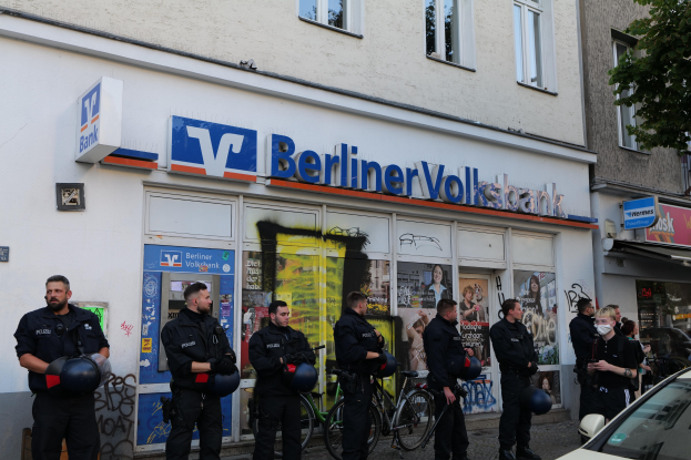 Polizisten vor einer Berliner Volksbank mit Helmen in den Händen und einem Auto auf der rechten Seite, einem Baum auf der linken Seite, Gebäude und Texttafeln im Hintergrund.