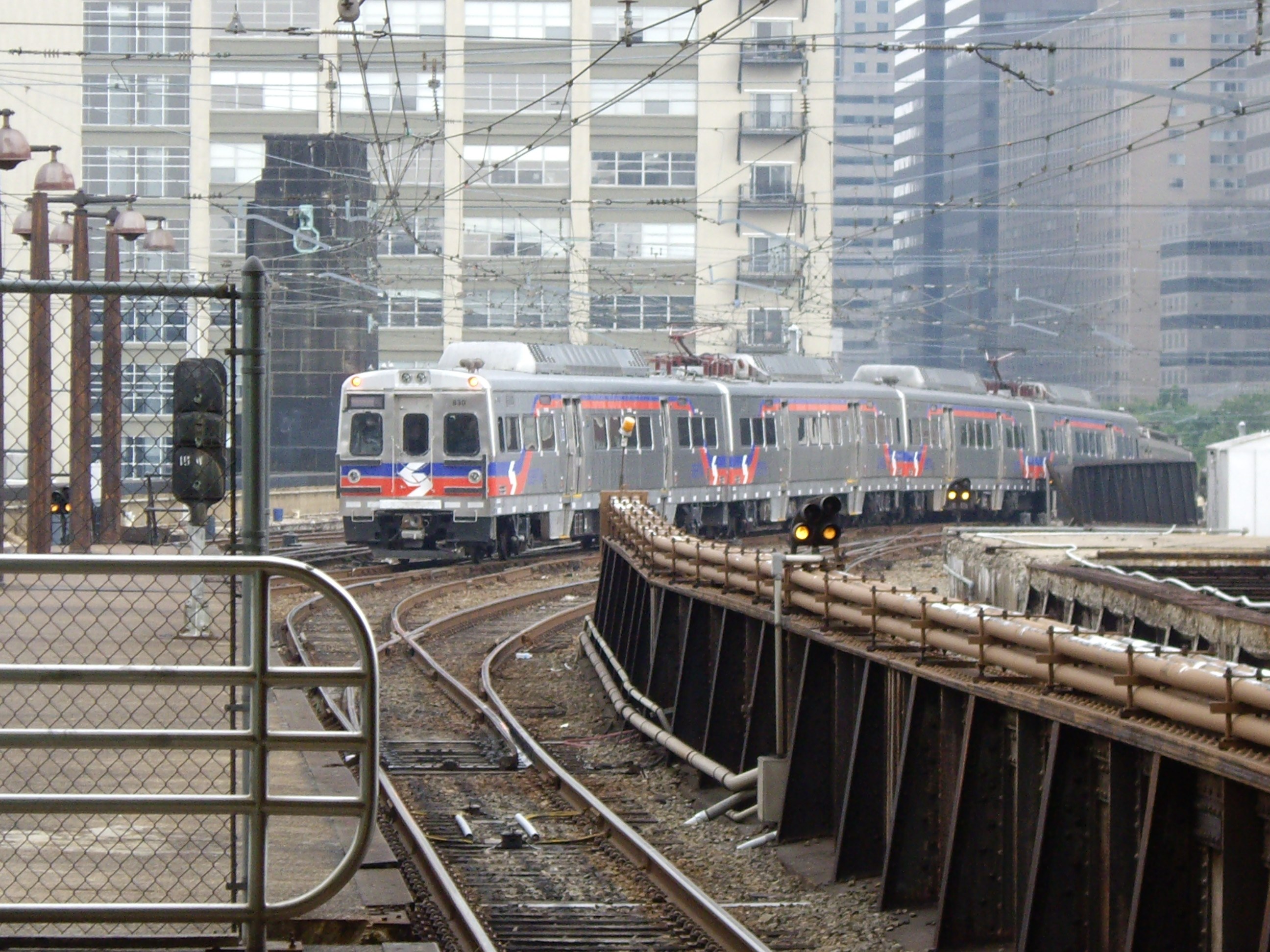 Nahverkehrszug fährt auf Schienen neben hohen Gebäuden mit städtischer Infrastruktur und Grünfläche im Hintergrund.