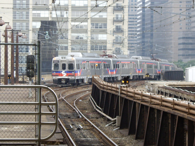 Nahverkehrszug fährt auf Schienen neben hohen Gebäuden mit städtischer Infrastruktur und Grünfläche im Hintergrund.