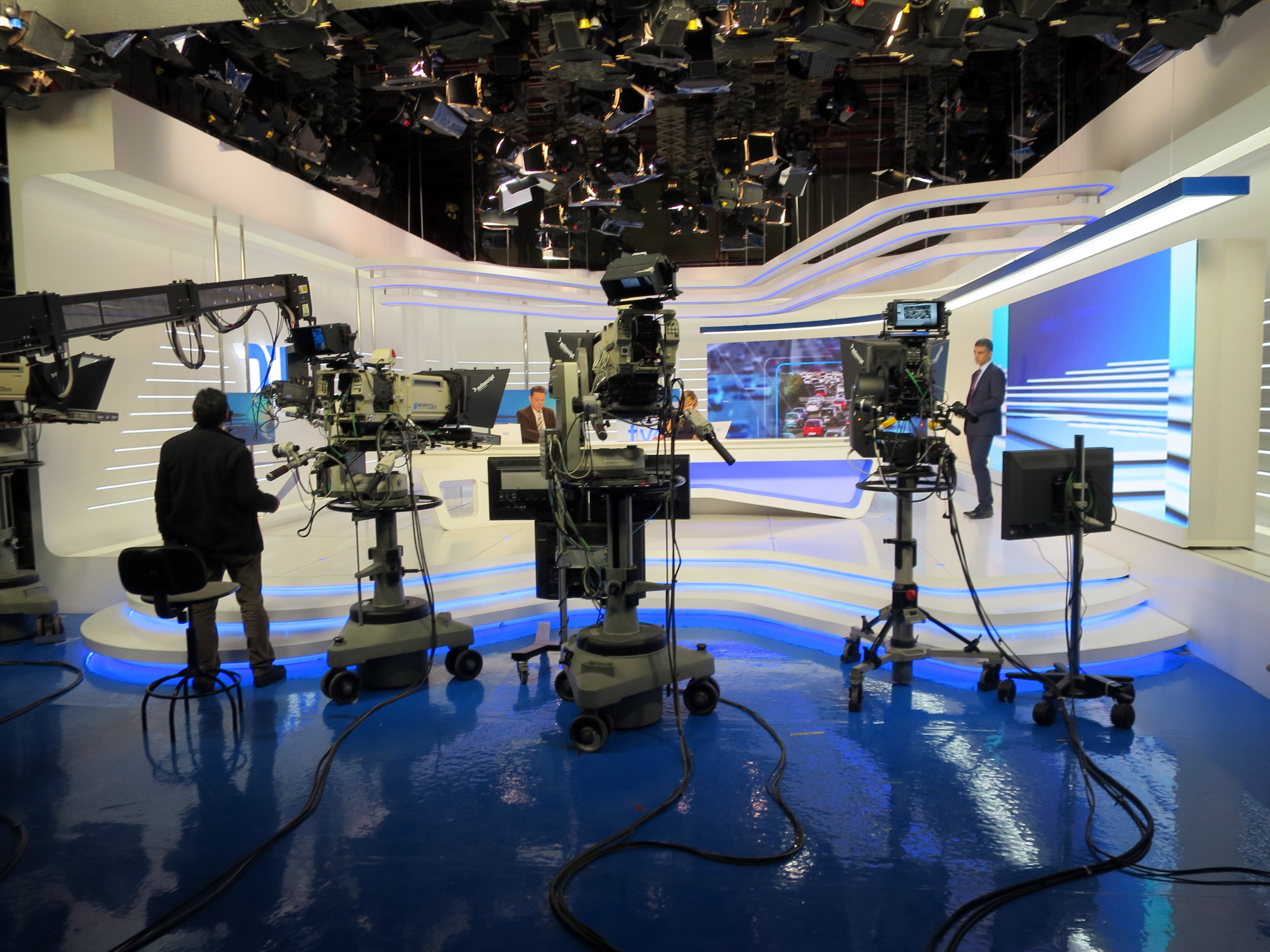 Ein Fernsehstudio mit mehreren Kameras auf dem Boden, einer Person im Vordergrund und einigen stehenden Personen im Hintergrund, mit Deckenleuchten für eine Livesendung.