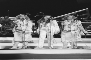 Drei Frauen singen in Mikrofone auf der Bühne mit Musikern hinter ihnen, in Schwarz-Weiß.