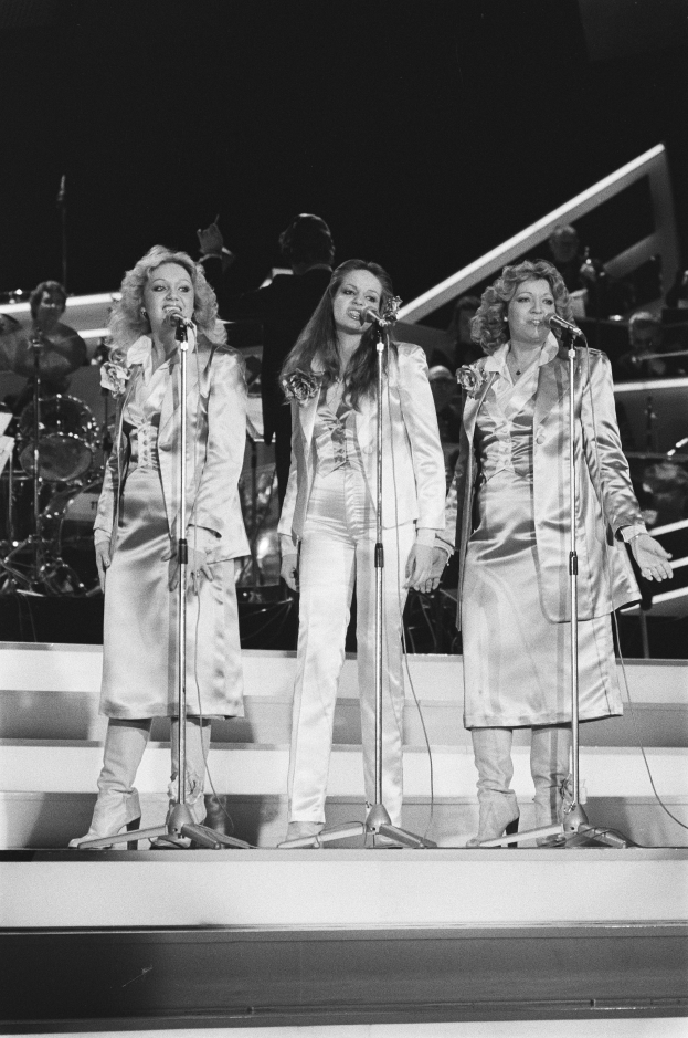 Drei Frauen singen in Mikrofone auf der Bühne mit Musikern hinter ihnen, in Schwarz-Weiß.