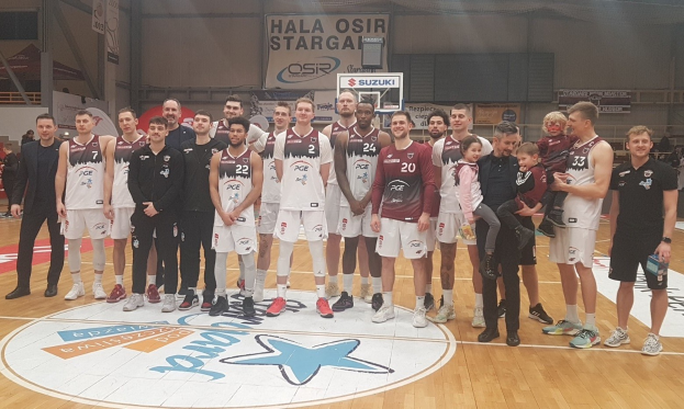Eine Gruppe von Männern und Kindern auf einem Basketballfeld mit Schildern, die "Hala Osir Starga" lesen, sitzende Zuschauer und ein Basketballkorb im Hintergrund.