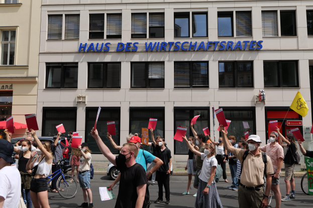 Eine Gruppe von Menschen demonstriert vor einem Gebäude, hält Fahnen und Schilder hoch, mit Fahrzeugen auf der Straße und einem Schild auf der rechten Seite.