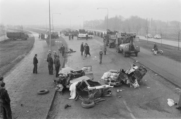 Eine schwarz-weiße Szene eines Autounfalls am Straßenrand mit mehreren Fahrzeugen, darunter ein Landfahrzeug, und einer Gruppe von Menschen drumherum, mit Laternenmasten, Bäumen und Himmel im Hintergrund.