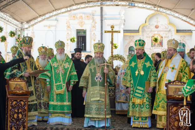 Eine Gruppe von Priestern steht vor einer Kirche während einer religiösen Zeremonie, wobei einer ein Buch und ein Mikrofon hält, während ein Kreuz, Blumen und ein Gebäude im Hintergrund sichtbar sind.