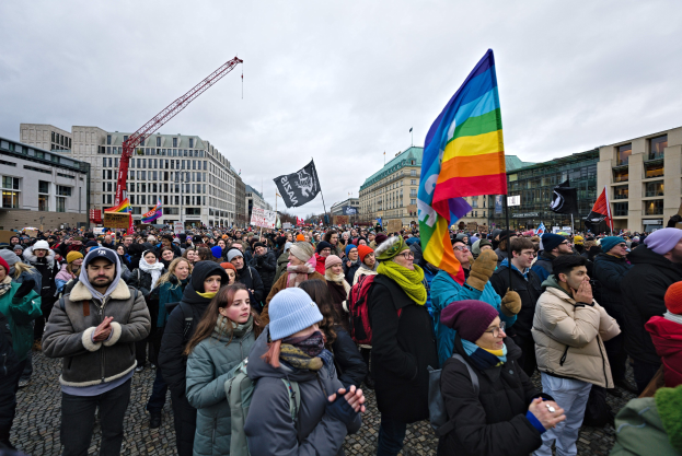 Große Gruppe von Menschen mit LGBTQ+-Rechten-Schildern und -Fahnen vor einem Gebäude mit einem Kran und einem bewölkten Himmel.