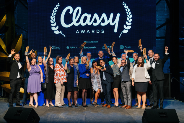 Eine Gruppe von Menschen auf einer Bühne, die lächeln und Auszeichnungen halten, mit Lautsprechern unten und einem Bildschirm im Hintergrund, der 'Classy Awards 2019' anzeigt.