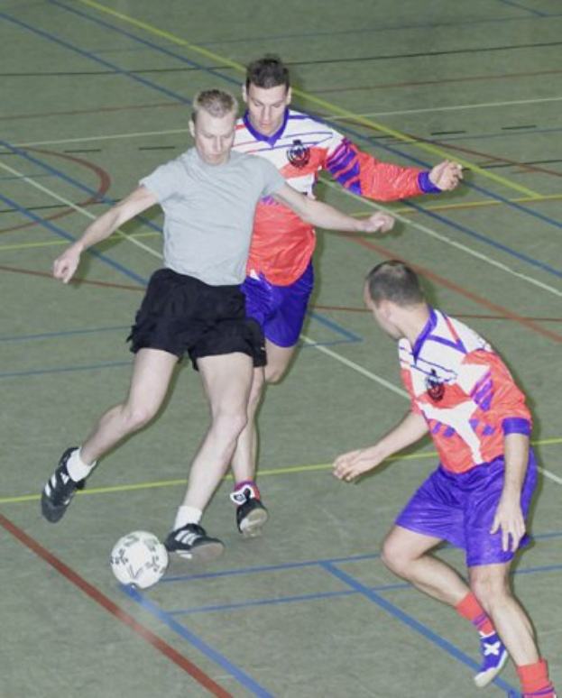 Drei Männer in bunten Trikots spielen Handball auf einem Platz, einer greift nach dem Ball in der Mitte.