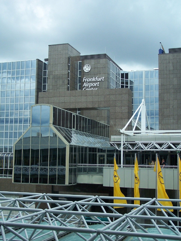 Großes gläsernes Gebäude des Frankfurter Flughafens in Frankfurt, Deutschland, mit Text an seiner Fassade, umgeben von gelben Flaggen und Metallpfosten unter einem teilweise bewölkten Himmel.