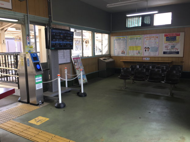 Ein belebter Bahnhofseingang mit Sitzgelegenheiten, einem Service-Schalter, Ticketautomaten, Kettenzäunen, Informationsanzeigen, Wandplakaten, Deckenbeleuchtung und Geländern.