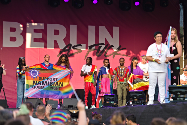 Gruppe von Menschen auf einer Bühne mit einem Banner, auf dem 'Berlin as Fuck' steht, mit Lautsprechern, Mikrofonen und einer feiernden Menge unten.