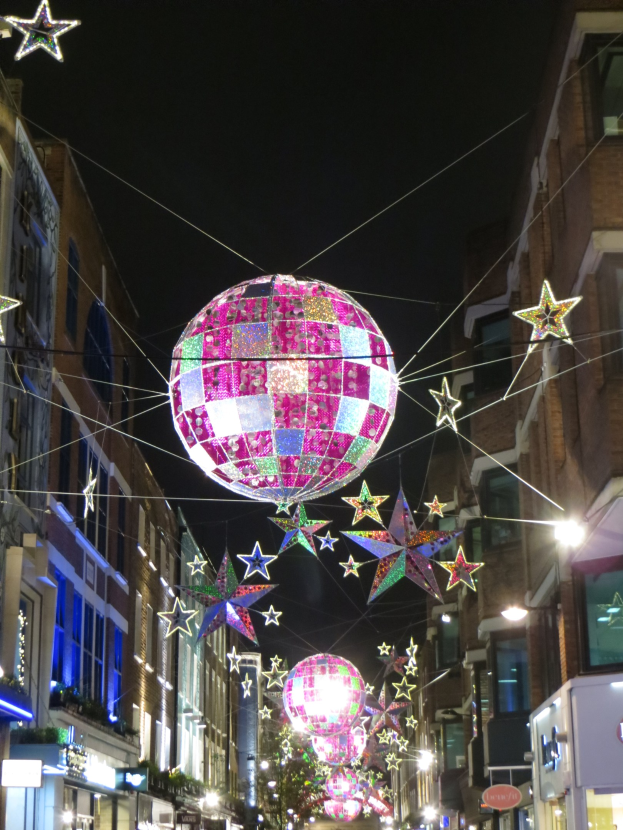 Eine nächtliche Stadtstraße mit zahlreichen Lichtern und Dekorationen, die einen großen Diskokugel von einem Gebäude hängen, schaffen eine festliche Atmosphäre.