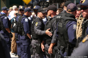 Eine Gruppe von Polizisten in Mützen, Taschen und Schutzbrillen, einige halten Waffen, steht zusammen auf einer Straße mit Gebäuden, Schildern und Polen im Hintergrund und Text unten im Bild.