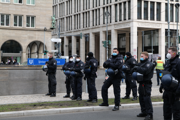 Eine Gruppe von Polizisten in schwarzen Uniformen und Masken steht vor einem gläsernen Gebäude mit Säulen in Berlin, Deutschland, einige halten Helme, mit Laternenmasten, Verkehrszeichen, Texttafeln, einer Statue, anderen Menschen und grasbewachsenem Boden im Hintergrund.