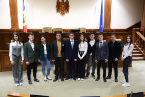 Gruppe von löchelnden Menschen, die vor einem Gerichtssaal mit Monitoren, Mikrofonen und anderen Gegenständen auf Tischen und einer hölzernen Plattform mit Flaggen und Stühlen im Hintergrund posieren.