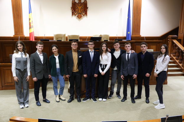 Gruppe von löchelnden Menschen, die vor einem Gerichtssaal mit Monitoren, Mikrofonen und anderen Gegenständen auf Tischen und einer hölzernen Plattform mit Flaggen und Stühlen im Hintergrund posieren.
