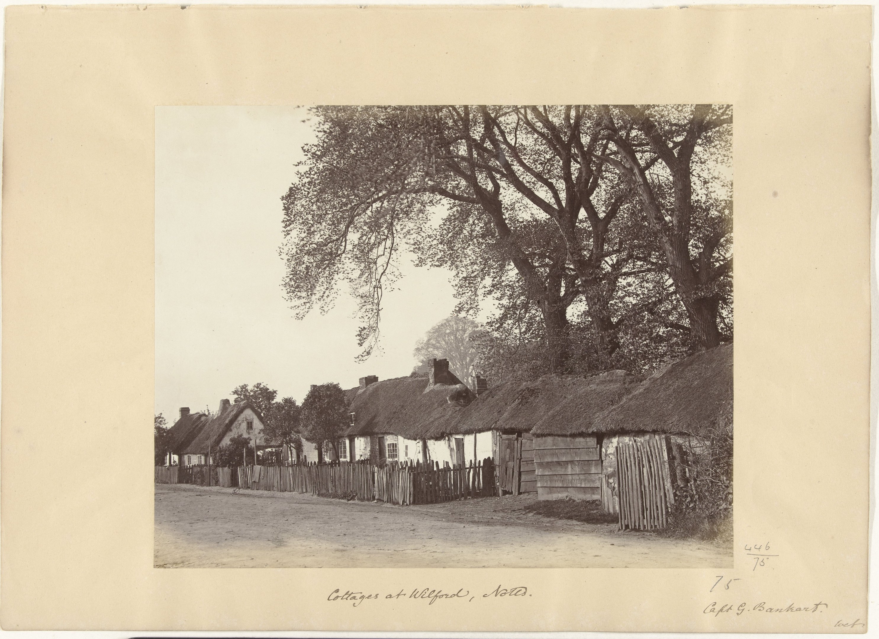 Schwarz-weiß-Foto eines Dorfes mit Reetdachhäusern, einer Straße, einem Zaun, Bäumen und einem Himmel im Hintergrund, mit Text unten.