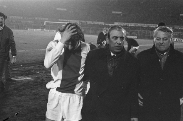 Schwarzes Bild einer Gruppe von Männern auf einem Fußballfeld mit einem Stadion im Hintergrund, die nach einem Sieg betrübt aussehen.