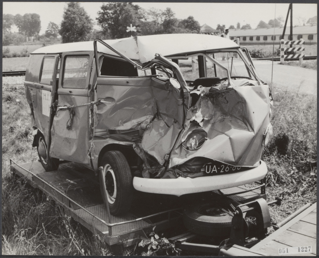 Ein Schwarz-Wei├č-Foto eines stark besch├Ądigten Autos auf einem H├Ąnger, umgeben von Gras und Pflanzen, mit B├Ąumen und einem Geb├Ąude im Hintergrund.