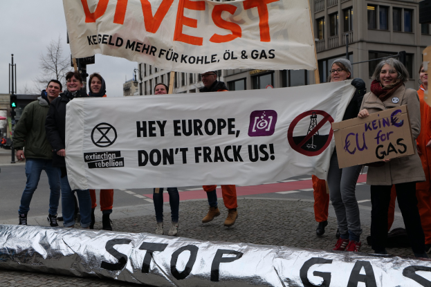 Eine Gruppe von Menschen auf einer Straße mit einem Banner "Stoppt Gas" im Protest gegen die Europäische Union, im Hintergrund Gebäude, Bäume, Pfähle, Verkehrsampeln und einen klaren blauen Himmel.