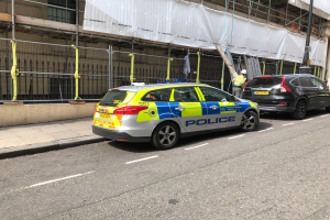 Ein Streifenwagen parkt an der Straßenseite neben einem Gebäude, zwei Helme tragende Personen stehen in der Nähe, ein Geländer und ein Pfahl sind im Vordergrund zu sehen, und ein weißes Tuch ist über das Gebäude drapiert, was auf eine Polizeiuntersuchung nach einem Messerangriff in London hindeutet.