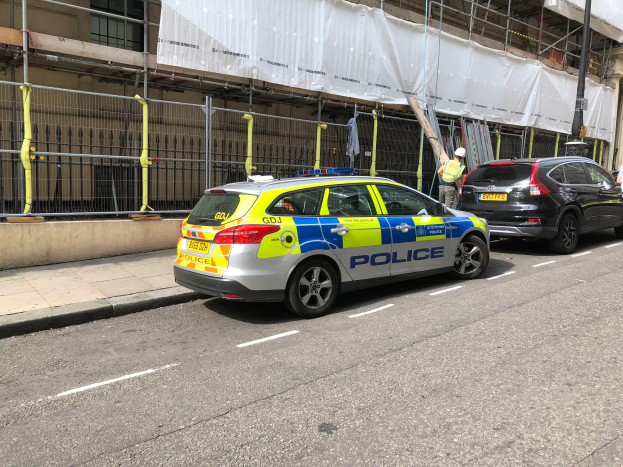 Ein Streifenwagen parkt an der Straßenseite neben einem Gebäude, zwei Helme tragende Personen stehen in der Nähe, ein Geländer und ein Pfahl sind im Vordergrund zu sehen, und ein weißes Tuch ist über das Gebäude drapiert, was auf eine Polizeiuntersuchung nach einem Messerangriff in London hindeutet.