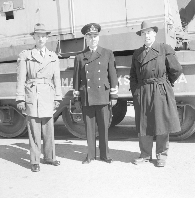 Drei Männer in Militäruniformen, Mänteln und Hüten stehen nebeneinander vor einem Zug, dargestellt in Schwarz-Weiß.