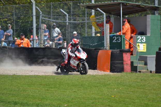 Ein Motorradfahrer in Helm und Handschuhen fährt über ein grasbewachsenes Feld, mit Zuschauern in Mützen und Schutzbrillen, einem eingezäunten Bereich, Treppen mit Geländern und einem nummerierten Schuppen im Hintergrund.