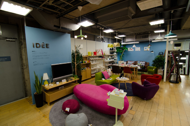 Eine Ladeneinrichtung mit Haushaltsgeräten und Möbeln, darunter ein Fernseher auf einem Tisch, eine pinkfarbene Couch, ein Sofa, zwei Stühle um einen kleinen Hocker, ein Esstisch mit Stühlen, ein großes Sofa im Hintergrund und verschiedene Gegenstände auf Regalen.
