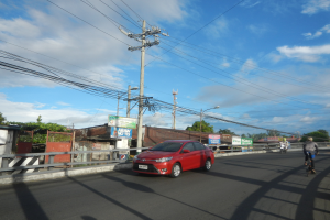 Ein rotes Auto fährt eine Straße entlang, gesäumt von Strommästen und -leitungen, mit einigen Radfahrern in der Nähe, Gebäuden, Bäumen und einem klaren blauen Himmel im Hintergrund.