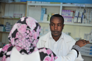 Eine Apothekerin in einem weißen Kittel spricht mit einer sitzenden Frau in einer Apotheke, mit Regalen voller Schachteln und anderen Gegenständen dahinter.