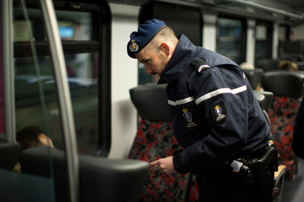 Ein Polizist in Uniform und Mütze steht in einem Zug und schaut auf sein Handy, umgeben von ein paar sitzenden Passagieren und einer weiteren Person auf der rechten Seite.