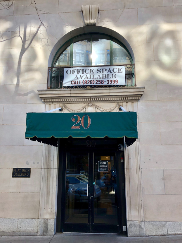 Glasstür mit grünem Vordach an einem Gebäude, mit einer Tafel, auf der 'Büror zu vermieten' steht, einem Geländer und einem Baum links.