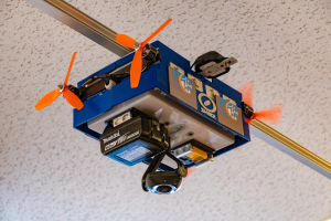 Ein kleiner blauer Drohne mit orangen Propellern fliegt in der Luft, befestigt an einer Metallstange an der Decke, wahrscheinlich für die Heimautomatisierung verwendet.