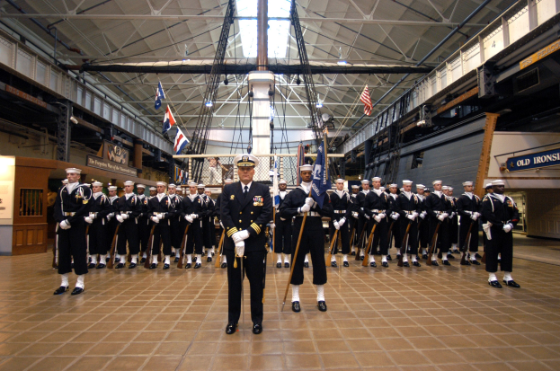 Eine Gruppe uniformierter Männer in einem großen Raum, die Mützen tragen und Gewehre halten, mit Flaggen und Informationsschildern im Hintergrund.