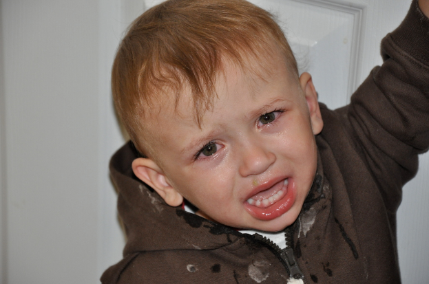 Ein kleiner Junge mit Jacke weint vor einer weißen Wand mit einer Tür im Hintergrund.