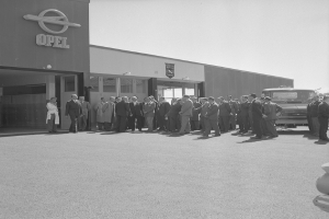 Eine Gruppe von Männern steht vor einer Opel-Vertretung mit einem Auto rechts daneben, in Schwarz-Weiß, mit Bäumen im Hintergrund.