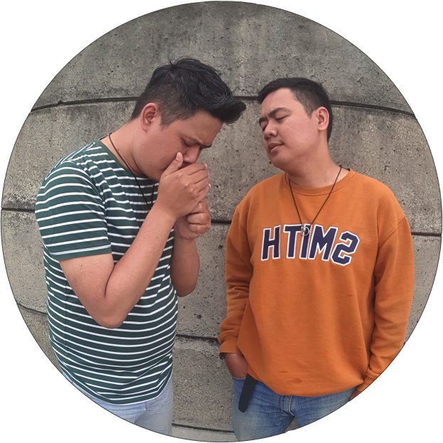 Zwei Männer stehen nebeneinander vor einer Wand, einer trägt ein orangefarbenes T-Shirt und blaue Jeans, der andere ein grün-weiß gestreiftes Hemd, beide wirken besorgt und sind in eine hitzige Diskussion vertieft.