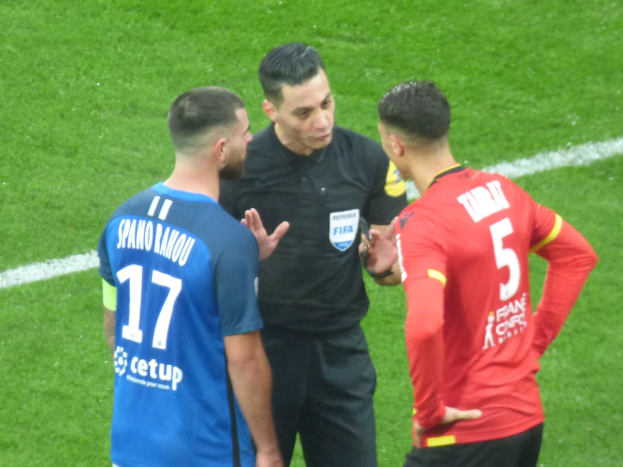 Schiedsrichter in schwarzem Hemd und Hose, der mit zwei Männern auf einem Fußballfeld spricht, einer in blauen und roten T-Shirts mit Text, beide tragen Brillen.