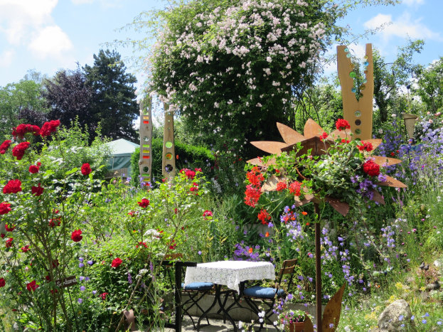 Ein Garten auf der Chelsea Flower Show mit bunten Blumen und Pflanzen, einem Tisch mit einer Decke und Stühlen, umgeben von Bäumen unter einem bewölkten Himmel.