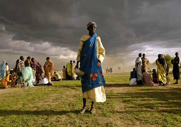 Eine Frau in einem blauen Kleid steht mit einem silbernen Gefäß in der Hand und wird von einer Gruppe von Frauen umgeben, die hinter ihr sitzen oder stehen, unter einem bewölkten Himmel mit Bäumen im Hintergrund.