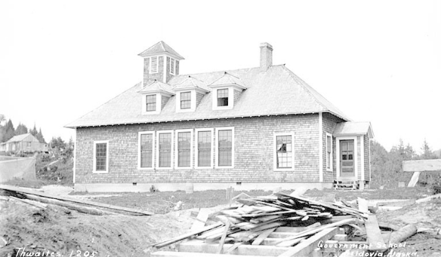Schwarzes und weißes Foto eines zweistöckigen Gebäudes mit Fenstern und Türen, umgeben von Bäumen und einem klaren Himmel, mit hölzernen Brettern unten, die "Regierungs-Schule, Thwaites, 1905" lesen.