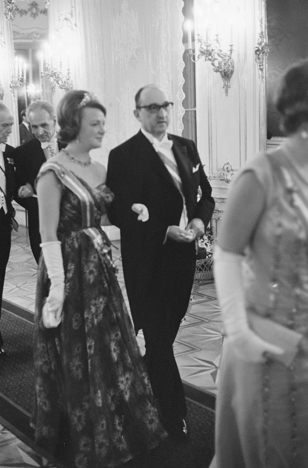 Ein Mann in einem schwarzen Blazer, weißem Hemd und schwarzen Hosen sowie eine Frau in einem eleganten Kleid gehen einen Flur entlang, umgeben von einer Gruppe von Menschen, die auf dem Boden stehen, während einige Wandlampen eine warme Atmosphäre schaffen.