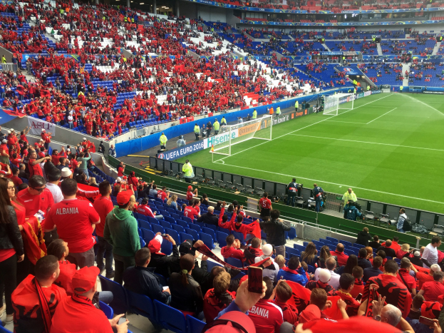 Ein volles Stadion mit Zuschauern, einige sitzen und einige stehen, mit Transparenten, einem Torpfosten und Deckenleuchten, während die Menge gespannt auf den Beginn eines Fußballspiels wartet.