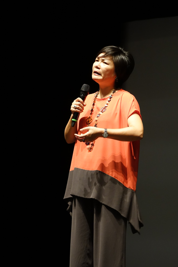 Frau auf einer hell erleuchteten Bühne mit Mikrofon, weißer Bluse und schwarzem Rock, mit Haaren im Dutt, spricht zu einem Publikum im Hintergrund.