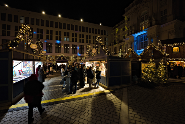 Ein lebendiger Weihnachtsmarkt in einer Stadt bei Nacht mit Menschen um beleuchtete Stände und geschmückte Gebäude im Hintergrund unter einem dunklen Himmel.